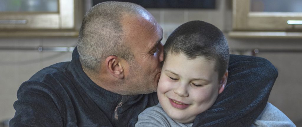 Dad and his son with autism sitting in kitchen at home. A man with short hair and bristle hugs his son and kisses him on the cheek.