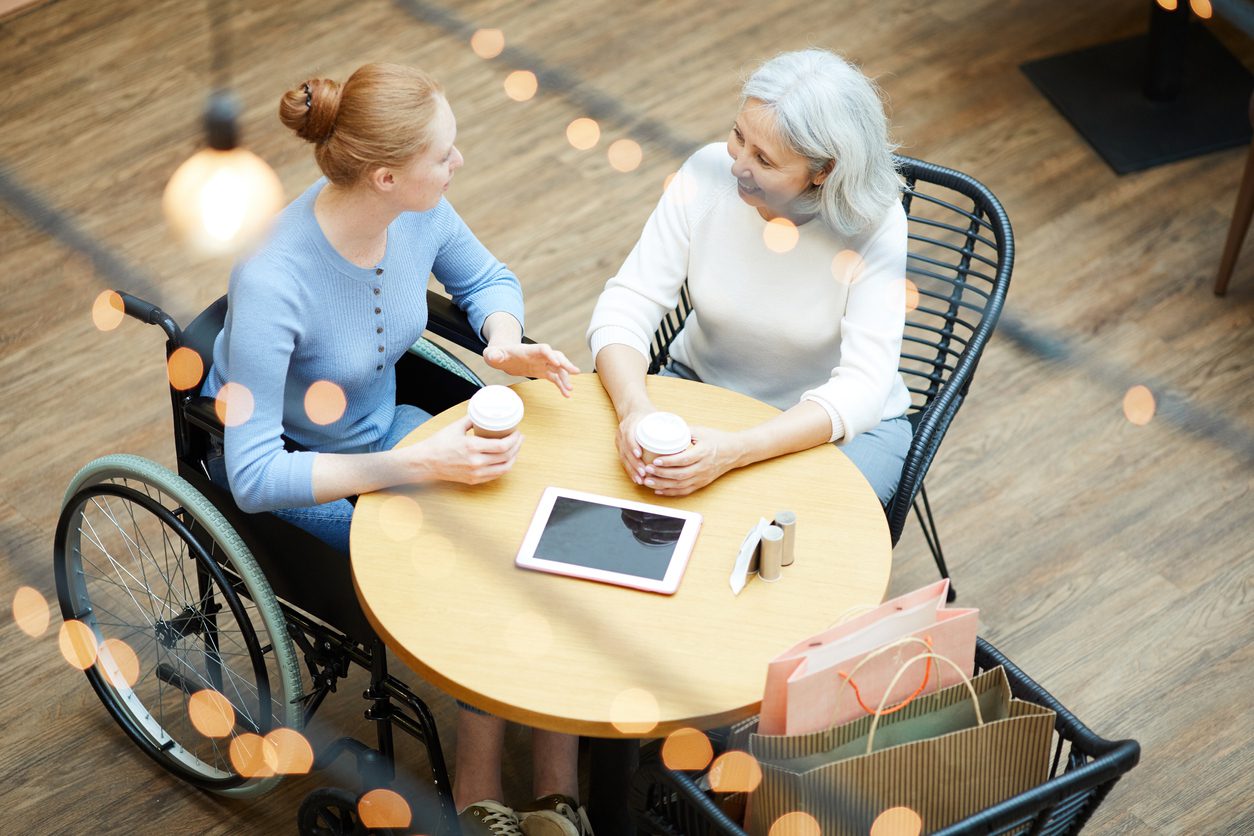 Young woman in wheelchair sitting at the table talking to her mother and drinking coffee at cafe after shopping in shopping mall